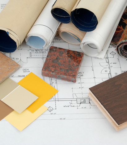 Rolled architectural blueprints and design materials, including paint swatches, tile samples, and wooden flooring, spread over detailed house floor plans, illustrating interior design and renovation planning.