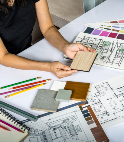 Interior designer working on a project, displaying wood samples and examining architectural drawings. The workspace includes color swatches, floor plans, and colored pencils, highlighting creative planning for interior design.